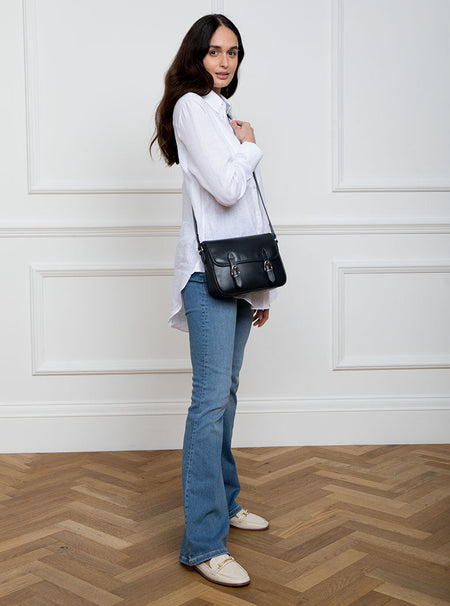 Woman wearing a large premium bridle butt navy leather crossbody Ceres hand bag that has been hand stitched in England with 2 front facing buckle straps and embossed with the Newmarket 875 horse logo on the back.